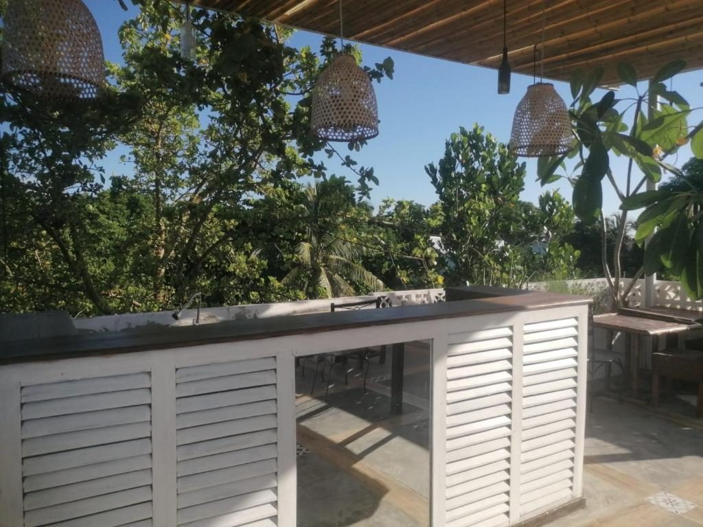 Cuisine d'été sur la terrasse couverte : comptoir blanc à persiennes, suspensions en vannerie et vue sur les arbres